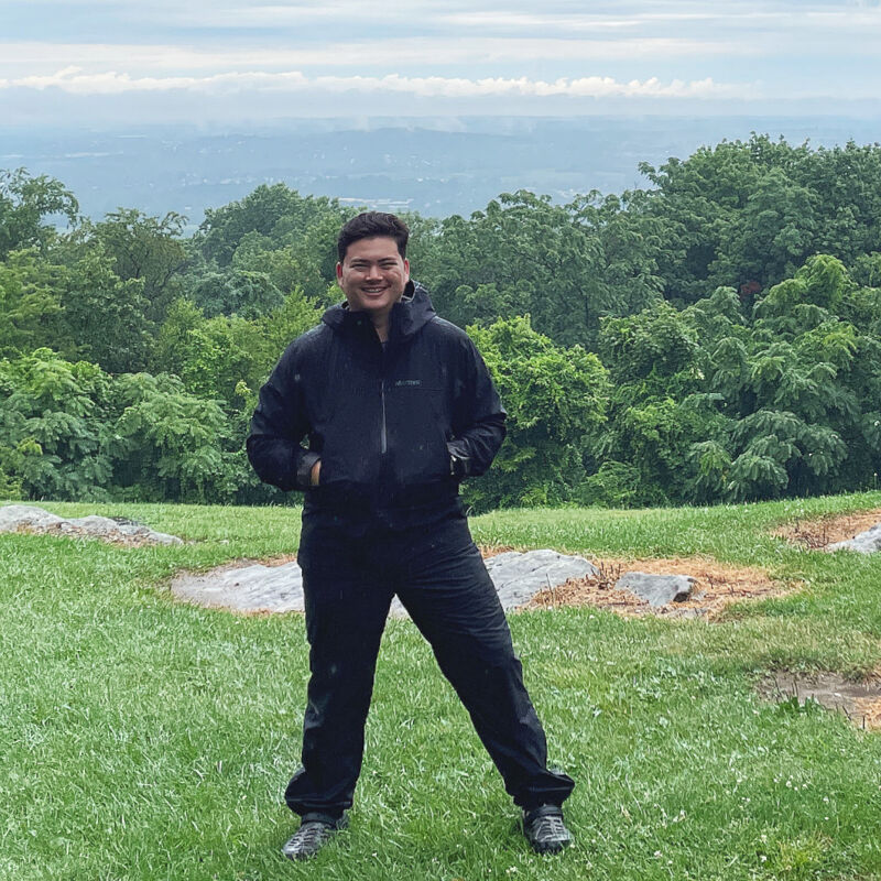 The image shows a man standing on a grassy hill with a scenic background. He is wearing a black jacket and pants. The background features lush green trees and a view of a distant mountain range under a cloudy sky. The man is smiling, suggesting a pleasant outdoor experience.